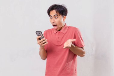 Yetişkin Güneydoğu Asyalı bir adam beyaz arka planda izole edilmiş, şok olmuş bir ifadeyle cep telefonuna bakıyor.