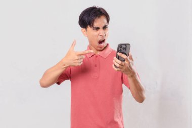Genç Asyalı adam öfkeyle cep telefonunu kullanıyor, sade arka planda izole edilmiş, ticari ve editörel kullanım için hayal kırıklığı ve duygu gösteriyor.