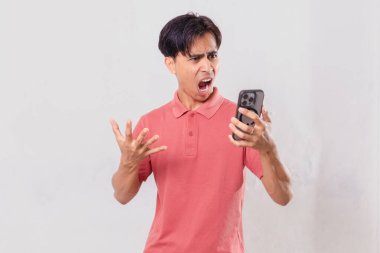 Genç Asyalı adam öfkeyle cep telefonunu kullanıyor, sade arka planda izole edilmiş, ticari ve editörel kullanım için hayal kırıklığı ve duygu gösteriyor.