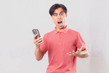 Genç Asyalı adam öfkeyle cep telefonunu kullanıyor, sade arka planda izole edilmiş, ticari ve editörel kullanım için hayal kırıklığı ve duygu gösteriyor.