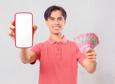 Neşeli, genç, Asyalı, boş akıllı telefon ekran modeli ve cep telefonu ve dijital ödeme konsepti için sade arka planda izole edilmiş para gösteren genç bir adam..