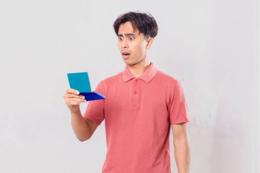 Banka hesap cüzdanı tutan genç bir adam, beyaz bir arka planda izole edilmiş hesap defterine bakarken şok olmuş ve kızgın bir yüz ifadesi takınıyor..