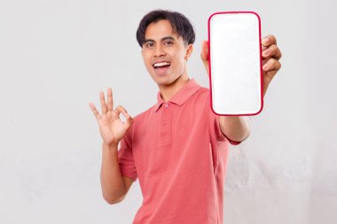 Heyecanlı genç Asyalı adam boş akıllı telefon ekran modeli gösteriyor ve mobil uygulama memnuniyeti için sade arka planda izole edilmiş ok işareti veriyor.