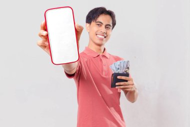 Neşeli, genç, Asyalı bir adam elinde para dolu bir cüzdan tutuyor ve dijital finans promosyonu için arka planda izole edilmiş boş akıllı telefon ekran modeli gösteriyor..
