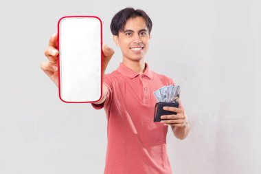 Neşeli, genç, Asyalı bir adam elinde para dolu bir cüzdan tutuyor ve dijital finans promosyonu için arka planda izole edilmiş boş akıllı telefon ekran modeli gösteriyor..