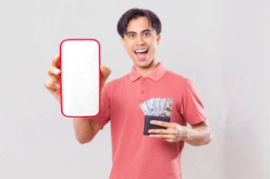 Neşeli, genç, Asyalı bir adam elinde para dolu bir cüzdan tutuyor ve dijital finans promosyonu için arka planda izole edilmiş boş akıllı telefon ekran modeli gösteriyor..