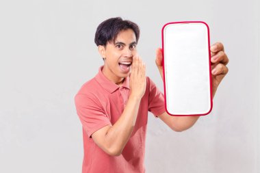 Heyecanlı genç Asyalı adam gizli terfi anlaşması için arka planda izole edilmiş fısıldayan hareketlerle boş akıllı telefon görüntüsü gösteriyor..