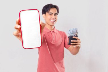 Neşeli, genç, Asyalı bir adam elinde para dolu bir cüzdan tutuyor ve dijital finans promosyonu için arka planda izole edilmiş boş akıllı telefon ekran modeli gösteriyor..