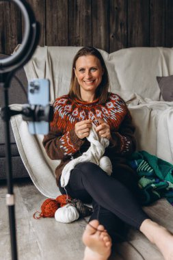 Samimi bir süveter giymiş gülümseyen bir kadın evden örgü örüp yayarken telefonuna el sallıyor. Kişisel video görüşmesi ya da video bloğu aracılığıyla sıcaklığı, yaratıcılığı ve rahatlığı paylaşmak. Yüksek kalite fotoğraf