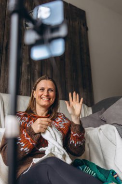 Samimi bir süveter giymiş gülümseyen bir kadın evden örgü örüp yayarken telefonuna el sallıyor. Kişisel video görüşmesi ya da video bloğu aracılığıyla sıcaklığı, yaratıcılığı ve rahatlığı paylaşmak. Yüksek kalite fotoğraf