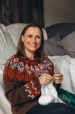 Kanepede oturan ve beyaz iplikle örgü ören bir kadın. Sıcak el yapımı hobi, evde rahatlama, sakin kış aktivitesi ve yaratıcı kişisel bakım konsepti. Yüksek kalite fotoğraf