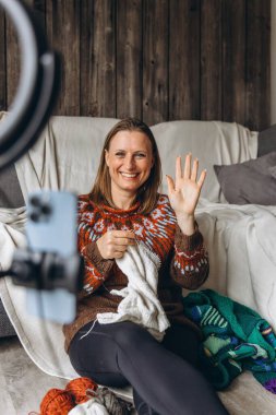 Samimi bir süveter giymiş gülümseyen bir kadın evden örgü örüp yayarken telefonuna el sallıyor. Kişisel video görüşmesi ya da video bloğu aracılığıyla sıcaklığı, yaratıcılığı ve rahatlığı paylaşmak. Yüksek kalite fotoğraf