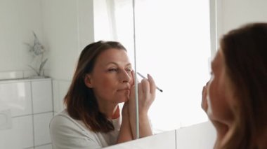 Orta yaşlı bir kadın banyo aynasının önünde fırçayla göz makyajı yapıyor. Günlük ev ortamında güzellik rutini ve kişisel bakım. Yüksek kaliteli FullHD görüntüler