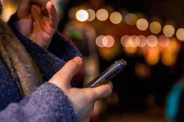 Bir gece pazarında, arka planda sıcak şenlik ışıklarının bulanık arkaplanına karşı akıllı telefonu tutan ellerin yakın çekimi. Seçici odak