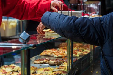 Bir müşteri, kışın markette çeşitli sıcak ve şenlikli sokak yemekleriyle dolu cam bir tezgahın arkasında kırmızı üniformalı bir satıcıdan taze pişmiş ekmek alır..