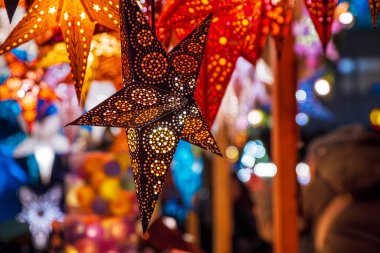 Canlı yıldız şeklinde kırmızı, turuncu, altın desenli fenerler Noel pazarında parlıyordu. Detaylı delinmiş tasarımlar ışıl ışıl ışık efektleri yaratır,