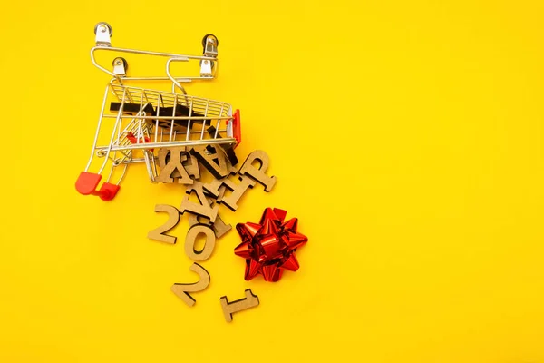 Feliz Año Nuevo 2021 se acerca. Las letras de madera cayeron de un carrito de comestibles con un lazo rojo sobre un fondo amarillo. Decoración festiva o concepto de postal. Vista superior y plana. Copiar espacio —  Fotos de Stock