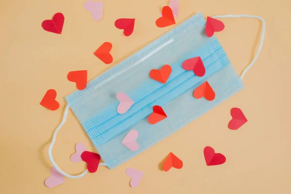 COVID-19. Face mask with paper cut out small hearts on an orange background. Medical masks to protect against viruses and pollution. Concept of celebrating valentine\'s day in a new reality.