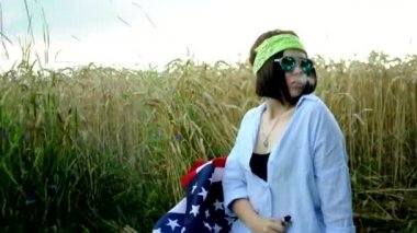 Bandana ve gözlüklü genç bir kız günbatımında bir tarlada elektronik sigara için popüler bir elektronik cihaz kullanıyor. Amerikan hippisi Amerikan bayrağının arka planında tüttürüyor..