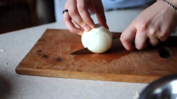 Chica corta la cebolla por la mitad para cortarla en trozos pequeños con un cuchillo afilado en una tabla de madera. El concepto de verduras frescas para cocinar en casa. — Vídeos de Stock