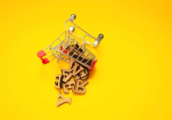 Letras de madera dispersas del alfabeto inglés de un carrito de compras en miniatura sobre un fondo amarillo, de cerca. Concepto de redacción y educación. Copiar espacio. —  Fotos de Stock