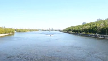 Tekne geniş bir nehirde yüzüyor, yukarıdan ön manzara. Turistli bir tekne, şehirde su turu yapıyor. Tekne nehir boyunca hızla ilerliyor. Köprü manzaralı..