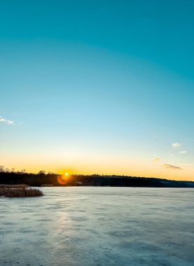 Sakin kış günbatımı donmuş gölün üzerine ılık bir ışık saçar, yukarısı açık mavi gökyüzü ve kıyıları kaplayan uzak ağaçlar, sakin bir atmosfer ve güzel doğal bir manzara yaratır.
