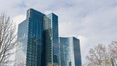 Parlak cam gökdelenler, ağaçlarla çevrili bulutlu bir gökyüzüne karşı yükseliyor, çağdaş mimari ve kentsel tasarım sergiliyorlar.