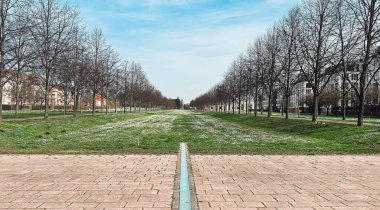 Serene park yolu, her iki tarafında düzgün sıralanmış ağaçlar barındırır, yemyeşil çimlere götürür, parlak mavi gökyüzünün altında huzurlu bir atmosfer yaratır, rahatlama ve keşiflere davetiye çıkarır.