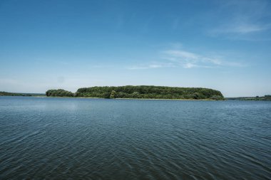 Huzurlu manzara, üzerinde berrak bir gökyüzü olan, sakin mavi sulara gömülü yemyeşil bir ada sergiliyor, doğa severler ve açık hava meraklıları için huzurlu bir atmosfer yaratıyor.