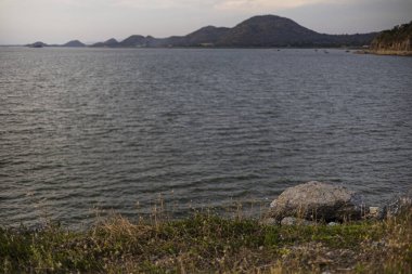 Grassy Slope By River at Dusk, sakin doğa kampanyası ya da huzurlu akşam belgeseli için, nehirdeki çimenli yamaçların, mor gökyüzünün sakin suları yansıttığı sakin bir fotoğraf..