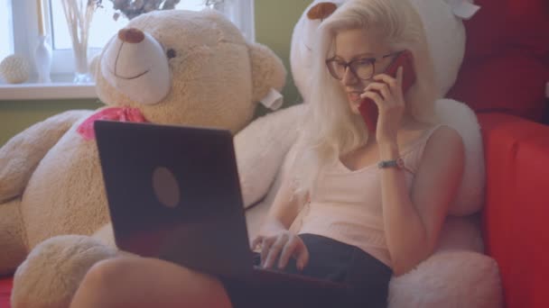 Блондинка играет и работает на ноутбуке и разговаривает по телефону — стоковое видео