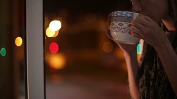 Девушка сидит на подоконнике, пьет горячий чай и смотрит на улицу. — стоковое видео