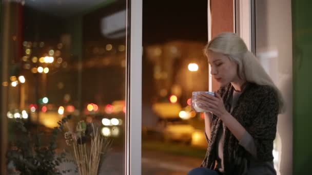 Girl sitting on the windowsill, drinking hot tea and looking out into the street — Stock Video