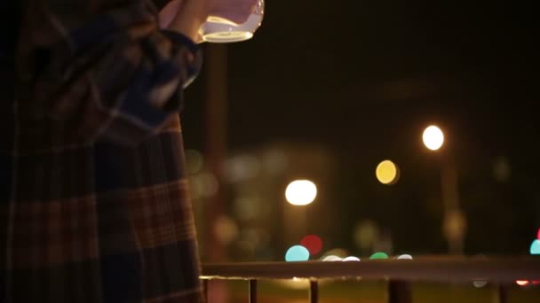 Una chica se para en el balcón con una taza de té, mirando el hermoso fondo urbano — Vídeos de Stock