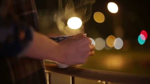 Una ragazza si trova sul balcone con una tazza di tè, guardando il bellissimo sfondo urbano — Video Stock