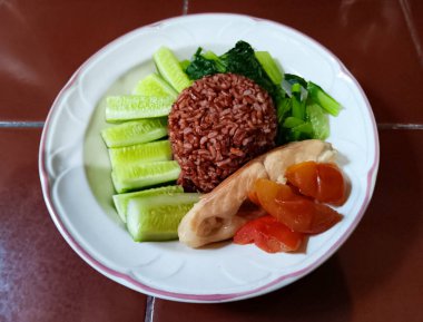 Healthy meal with red rice, sliced cucumbers, boiled bok choy, sliced boiled red tilapia with tomatoes on white plate isolated on terracotta tile countertop.