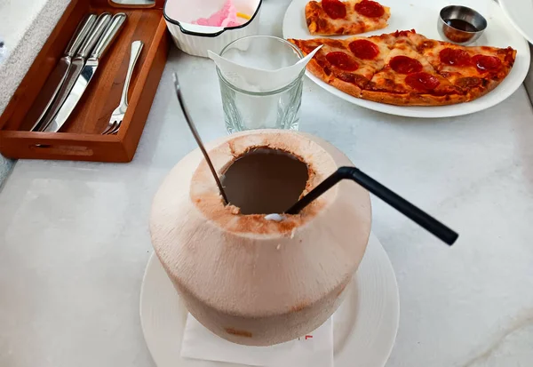 Siyah kaşık ve kaşık ile servis edilen taze bir hindistan cevizi. Restorandaki beyaz mermer masada izole edilmiş pepperonili pizza.. 