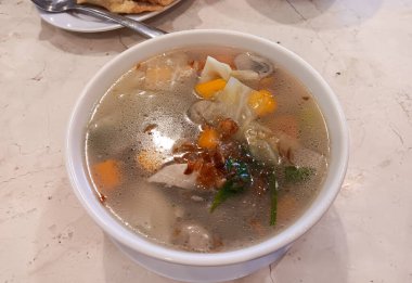 Endonezya Çin yemeği, sebzeli berrak tavuk çorbası: havuç, lahana, mantar, patates, domates üzerine kızarmış arpacık soğanı, mermer masada izole edilmiş beyaz bir kase..