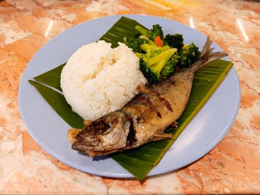 Geleneksel Endonezya yemeği, kızarmış uskumru balığı, yanında beyaz pilav ve muz yaprağı ve mermer masada izole edilmiş mavi tabakta sunulan karıştırılmış brokoli..