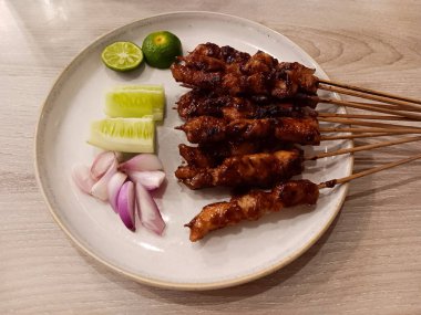 Close up of Indonesian chicken satay, skewers of grilled chicken commonly known as sate ayam served with sliced red onions, cucumber pieces and lime in a white plate isolated on wooden background.
