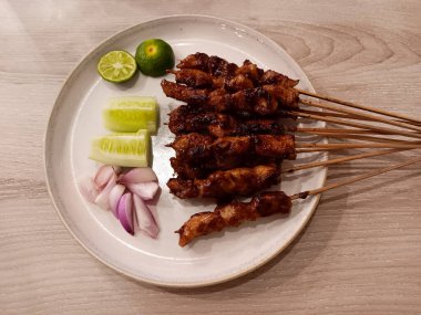 Endonezya usulü tavuk satay kapağı, ızgara tavuk şişleri genellikle sate ayam olarak bilinir dilimlenmiş kırmızı soğan, salatalık parçaları ve ahşap arka planda izole edilmiş beyaz bir tabakta limon ile servis edilir..