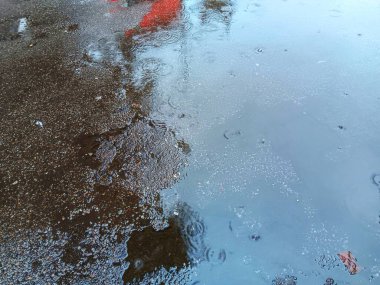 Soyut doku görüntüsünü kapat. Yağmur damlaları asfalt yolda büyük bir su birikintisi ve yağmurlu bir günde gökyüzünün yansıması ile dalgalanır..