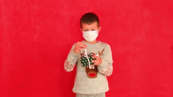 Un niño con una máscara médica y un suéter de Año Nuevo sobre un fondo rojo. — Vídeo de stock