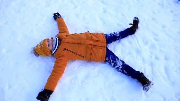 Un jeune garçon vêtu d'une veste jaune se trouve dans la neige et fait un ange de la neige. — Video