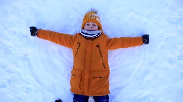 Um menino de casaco amarelo jaz na neve e faz um anjo da neve. — Vídeo de Stock