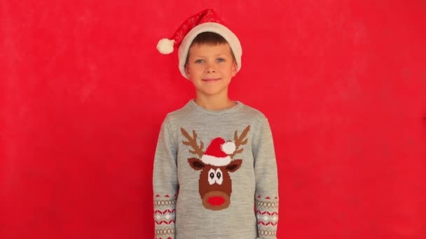 Niño en una gorra de Año Nuevo y suéter sobre un fondo rojo. — Vídeo de stock