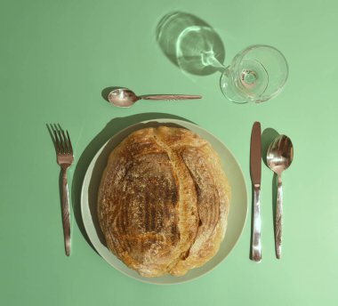 Tabakta servis edilen esnaf ekmeği ve pastel yeşil arka planda çatal bıçak. Çağdaş natürmort dengeyi, ışığı ve günlük yiyeceklerin şiirselliğini keşfediyor. Minimalist modern kahvaltı konsepti.