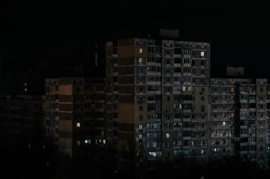 Karartma sırasında 2025 yılında bir şehri karanlık kaplar. Apartman binaları gece gökyüzüne karşı siluet vaziyette duruyor. Bazı pencerelerde sadece birkaç loş ışık görünüyor.