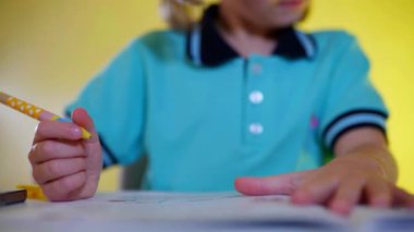 Açık renk saçlı, tıknaz gözlüklü, beyaz bir masada ödevlerine odaklanmış bir çocuk. Parlak mavi üniformalı genç bir öğrenci, parlak sarı bir arka planla aydınlatılmış bir çalışma kitabına özenle yazıyor..